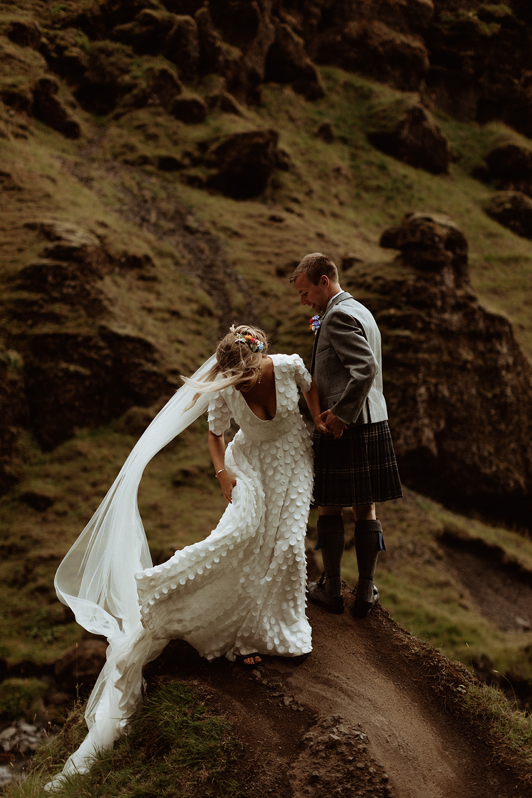 Bride and groom standing on a rock at Kvernufoss waterfall, with the wind lifting the bride’s veil, capturing the raw beauty of their Iceland elopement.