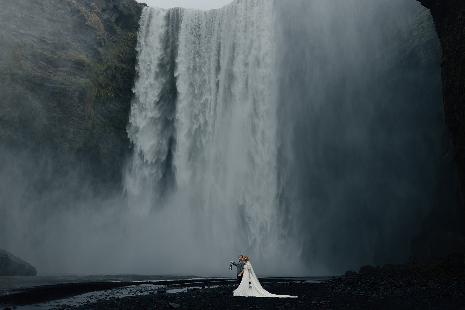 Bride and groom standing in front of Skógafoss waterfall, surrounded by mist, capturing the raw beauty of their Iceland elopement.