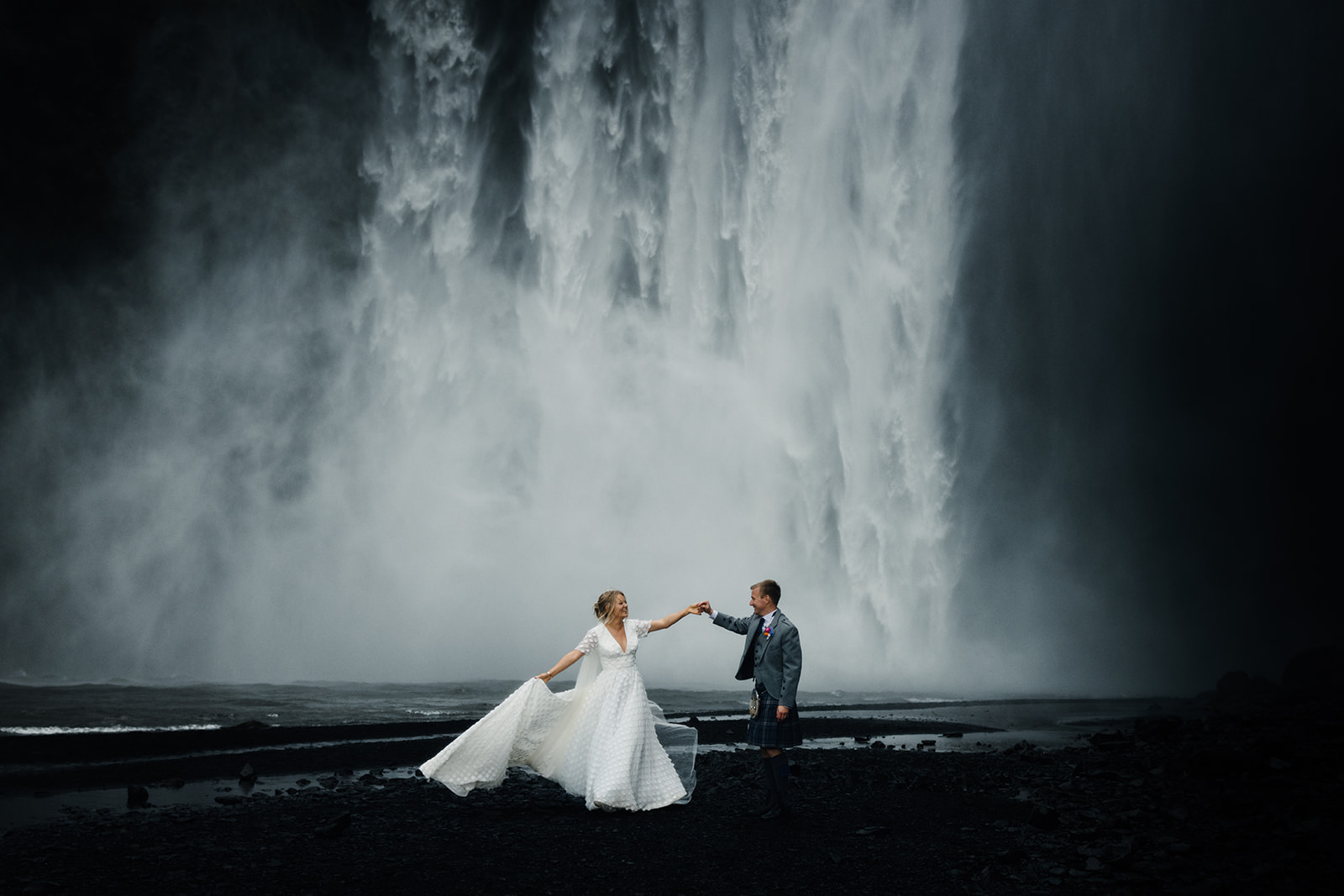 Newlywed couple dancing under the towering Skógafoss waterfall, mist swirling around them as they embrace the moment.