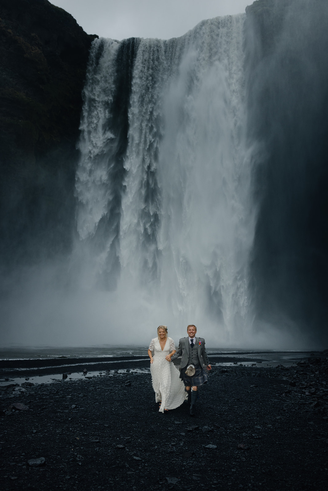 Bride and groom running towards the camera in front of Skógafoss waterfall, capturing the joy and excitement of their Iceland elopement