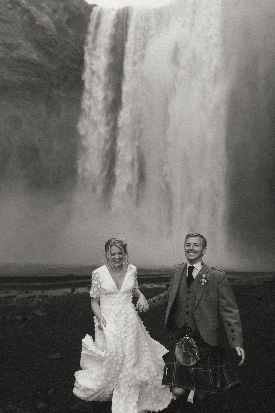 Black and white image of bride and groom running towards the camera in front of Skógafoss waterfall, capturing the emotion and raw beauty of their Iceland elopement