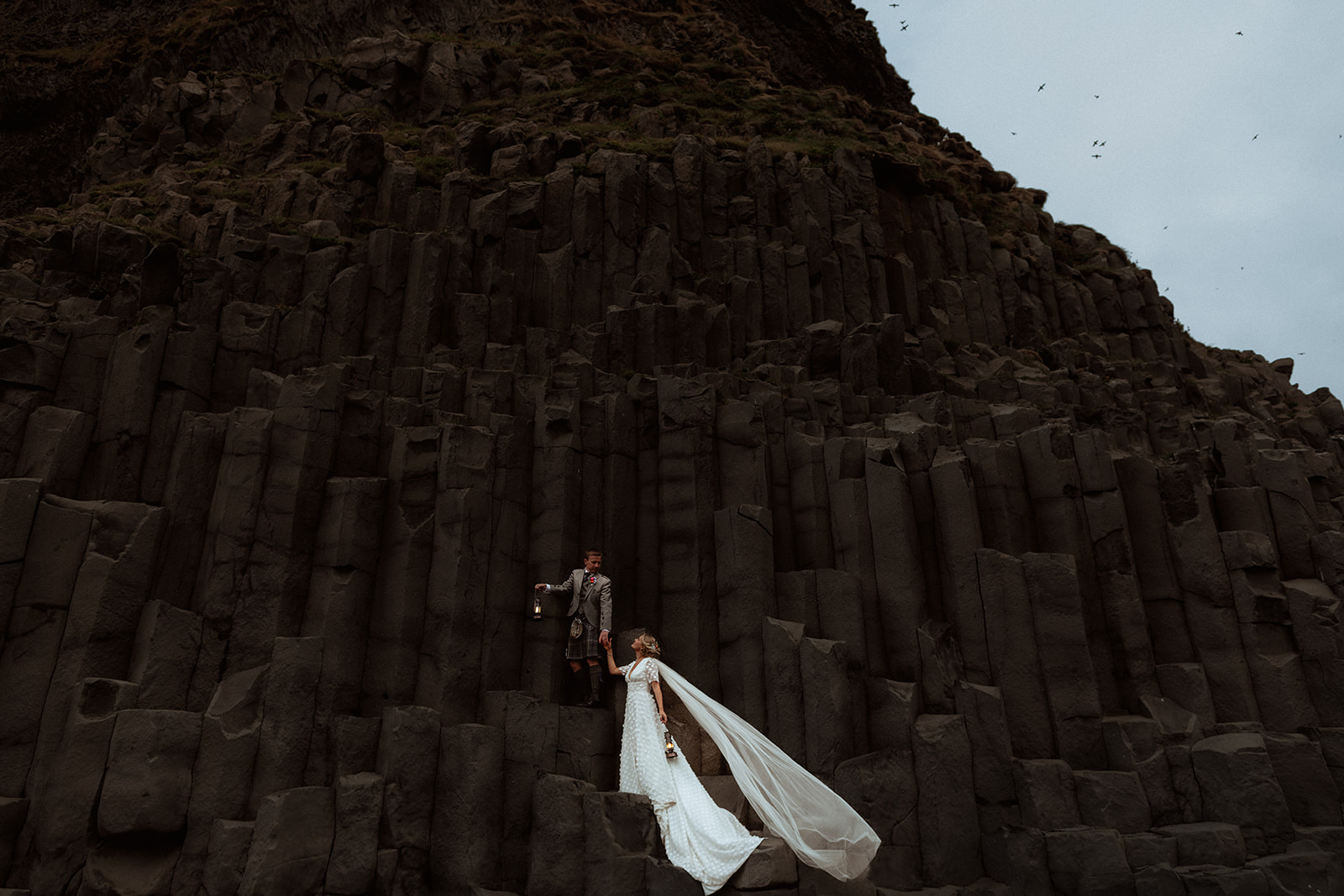 Amy and Tom walk hand-in-hand along Reynisfjara Beach, the iconic basalt columns creating a striking backdrop for their elopement adventure