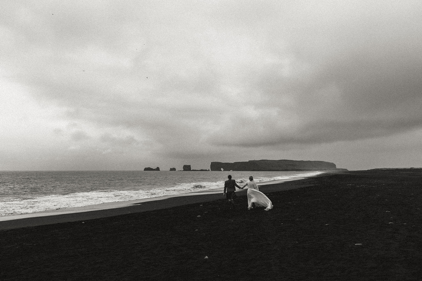 The couple joyfully runs together on the black sand of Reynisfjara Beach, with Iceland's iconic sea stacks behind them.