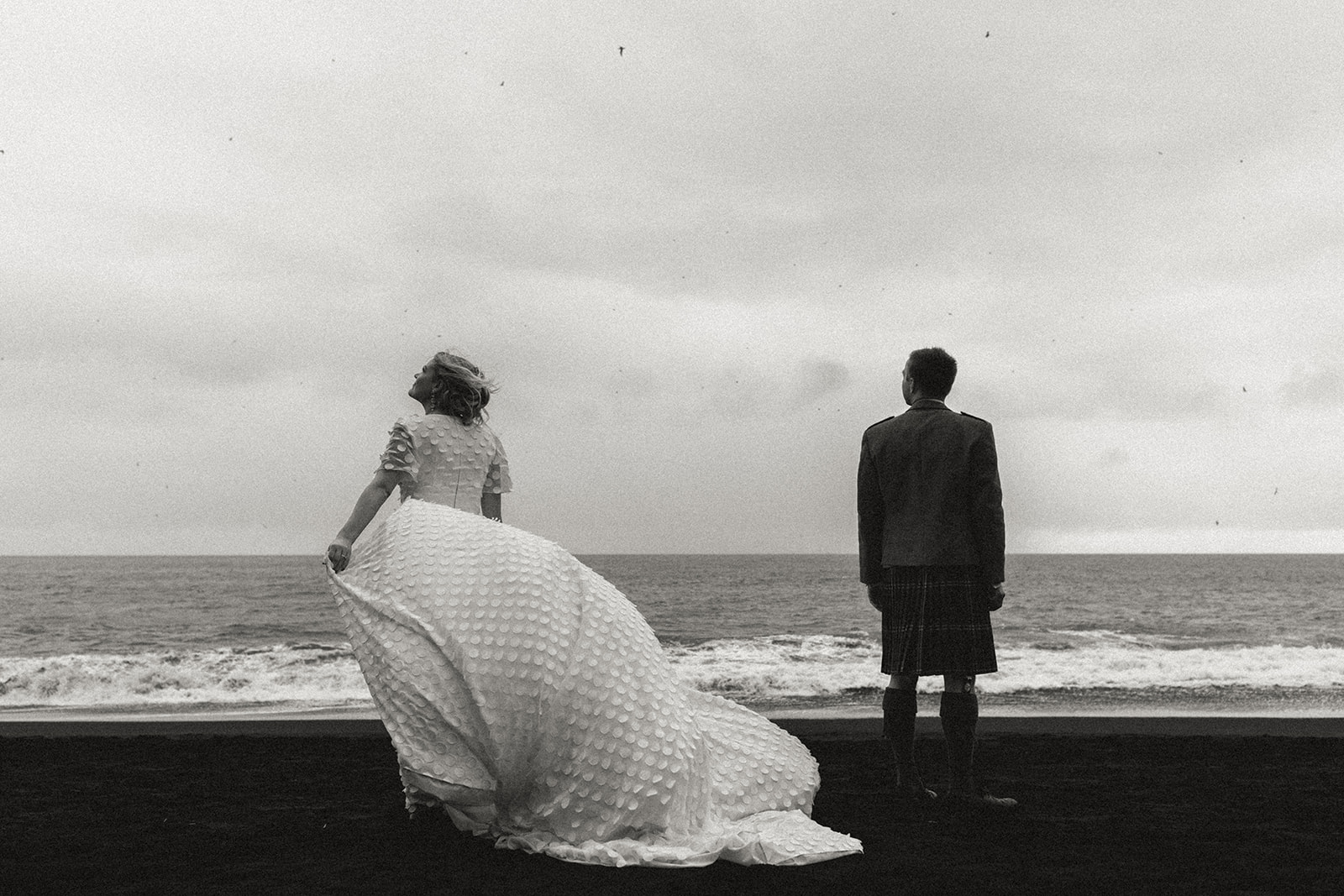 Artistic shot of newlyweds standing together on the black sand beach at Reynisfjara, with dramatic cliffs and ocean waves in the background.