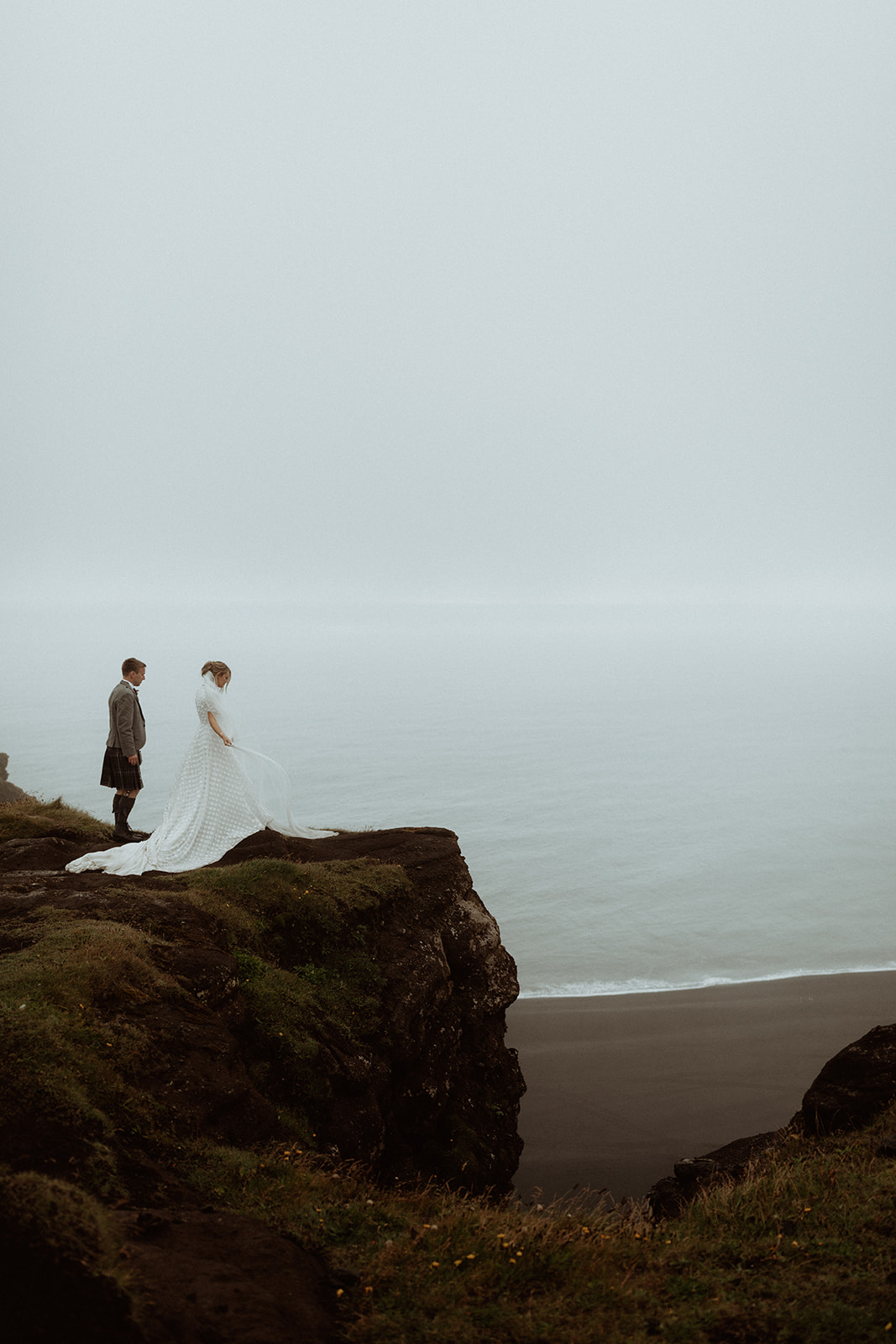 Bride and groom overlook Dyrhólaey in Iceland, taking in the sweeping coastal views during their elopement adventure.