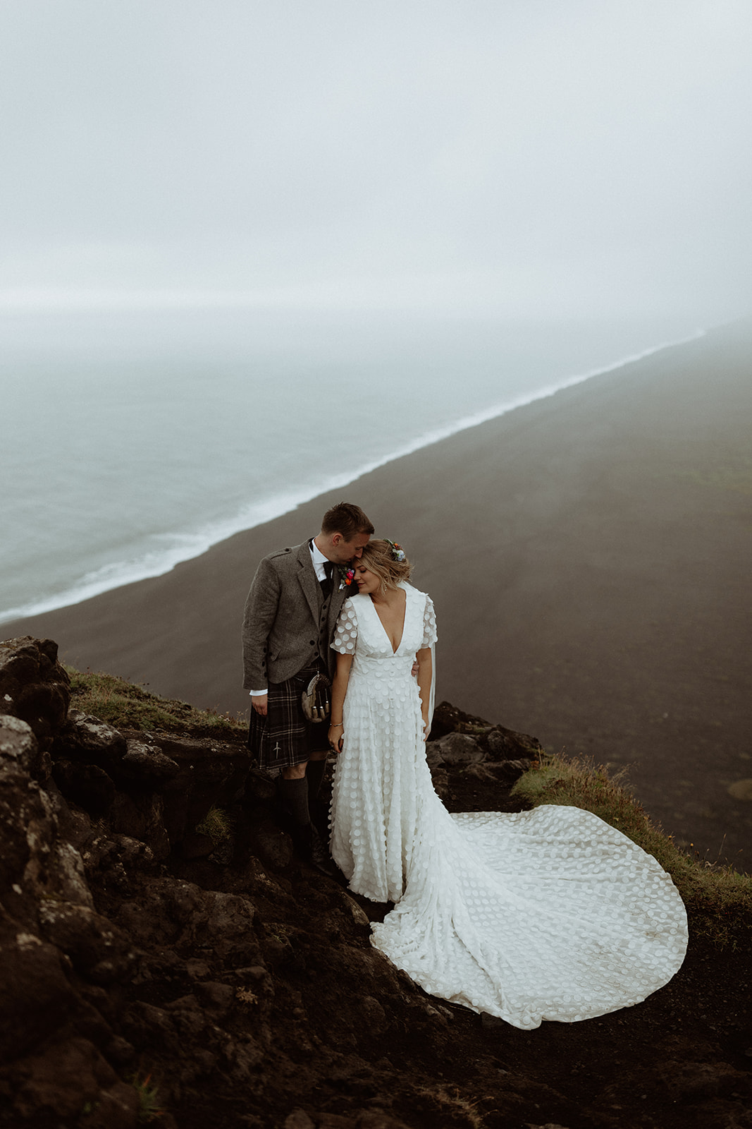 Bride and groom embrace the beauty of Dyrhólaey, the wind catching her veil as they enjoy the panoramic views during their elopement.