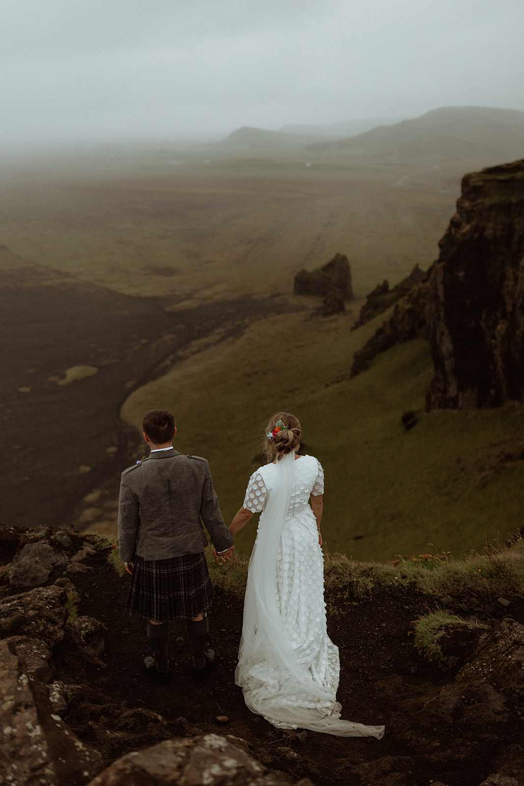A wide-angle shot of the bride and groom standing at Dyrhólaey, overlooking the rugged coastline as part of their Iceland elopement.