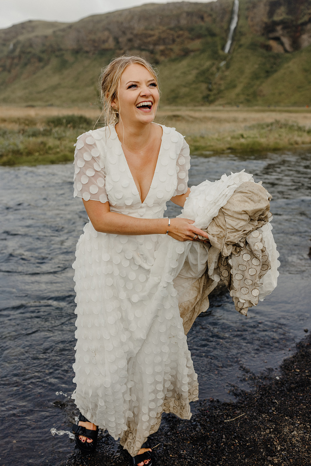 Bride laughing and lifting her muddy dress while washing her feet in a stream during her Iceland elopement.
