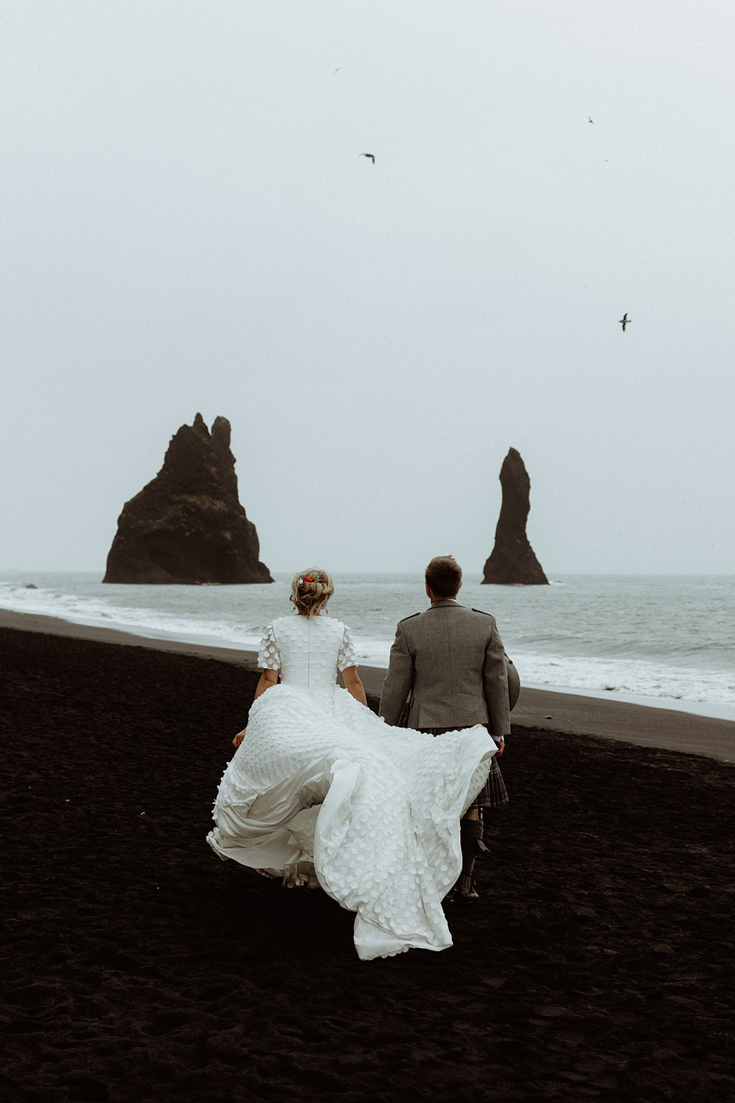 Bride and groom running hand-in-hand across the black sand beach at Reynisfjara, Iceland, with towering sea stacks in the background.