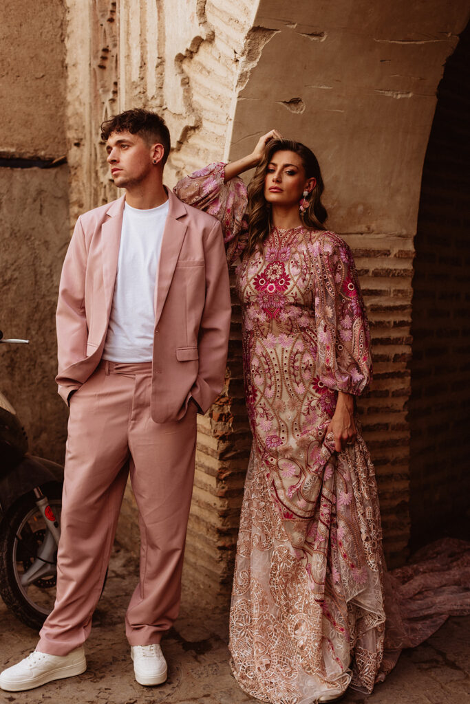 Bride and groom in coordinated pink and patterned outfits against a Marrakesh wall