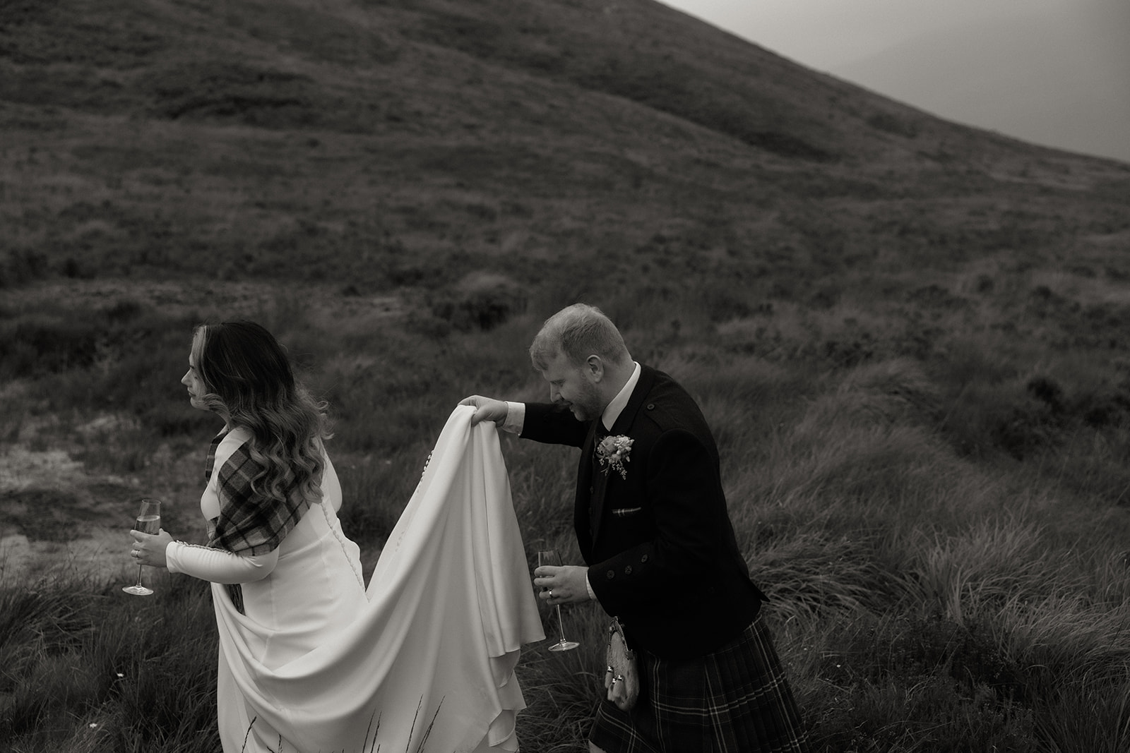 Groom lifting bride’s dress while walking by loch during Isle of Arran elopement.