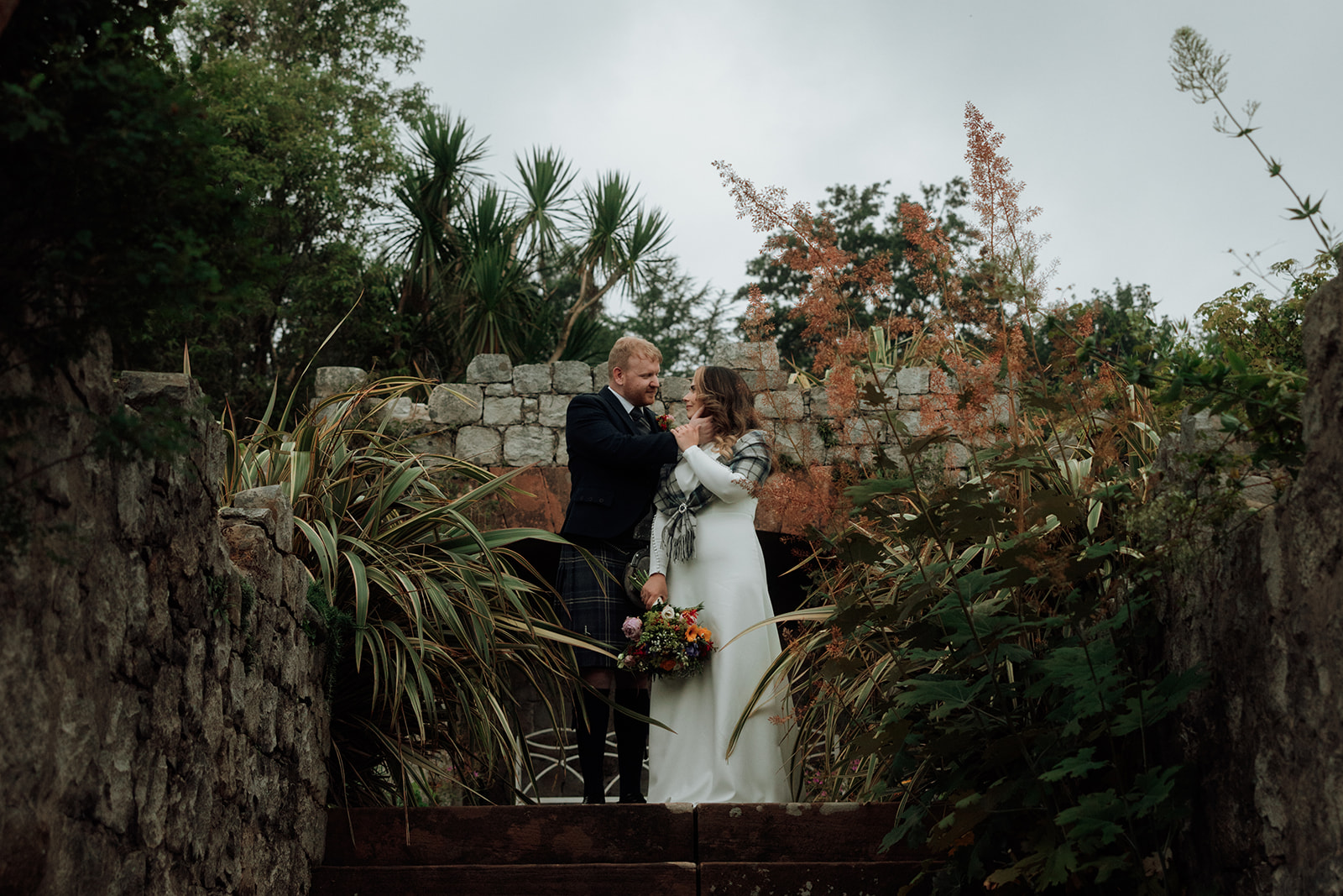 Bride and groom standing by stone wall with plants during Isle of Arran elopement.