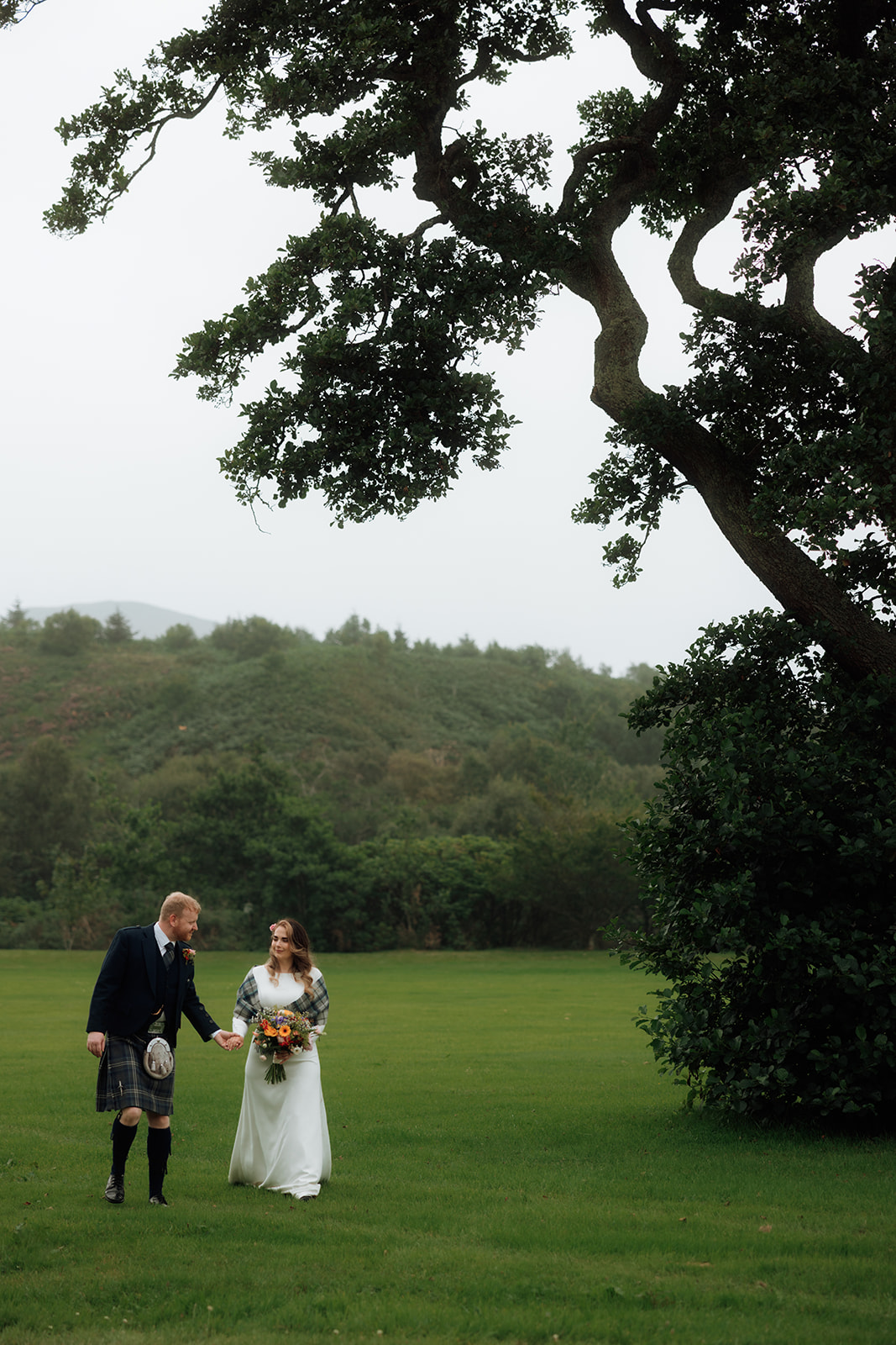 Bride and groom walking across lawn with large tree behind them on Isle of Arran.