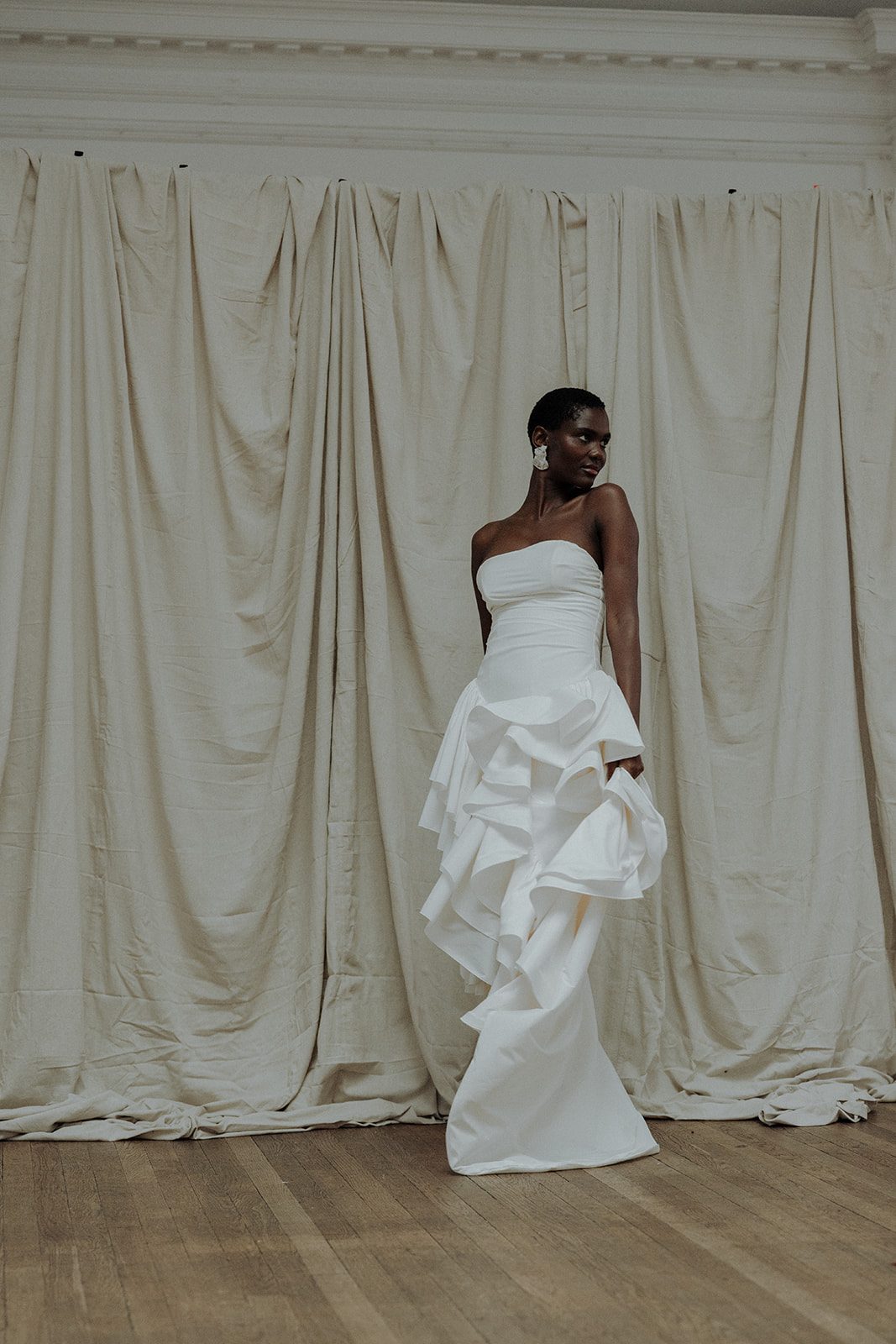 Bride in strapless couture gown standing tall against curtain backdrop, full-length editorial portrait.