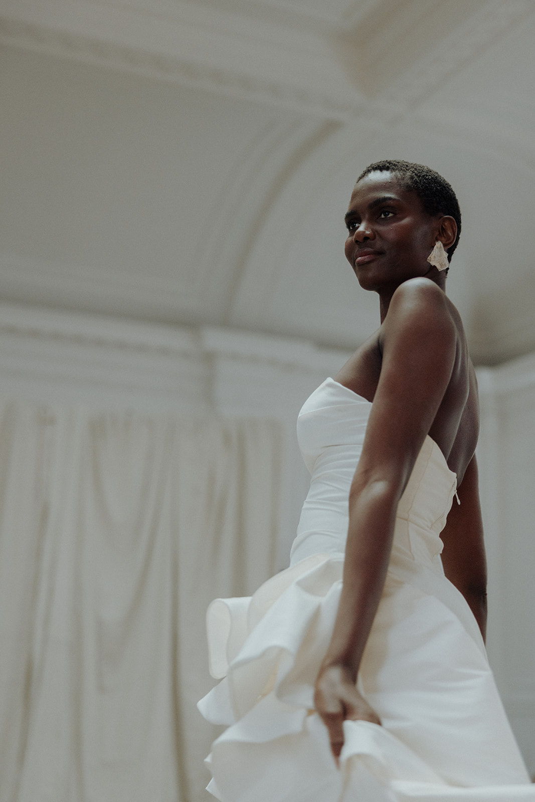 Bride in strapless couture gown, three-quarter profile with curtain backdrop, editorial bridal photograph.