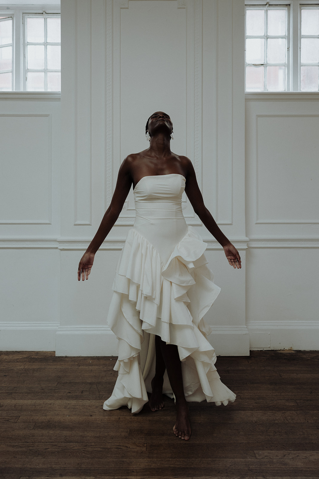 Bride in strapless couture gown standing full-length against white wall, editorial bridal photograph.