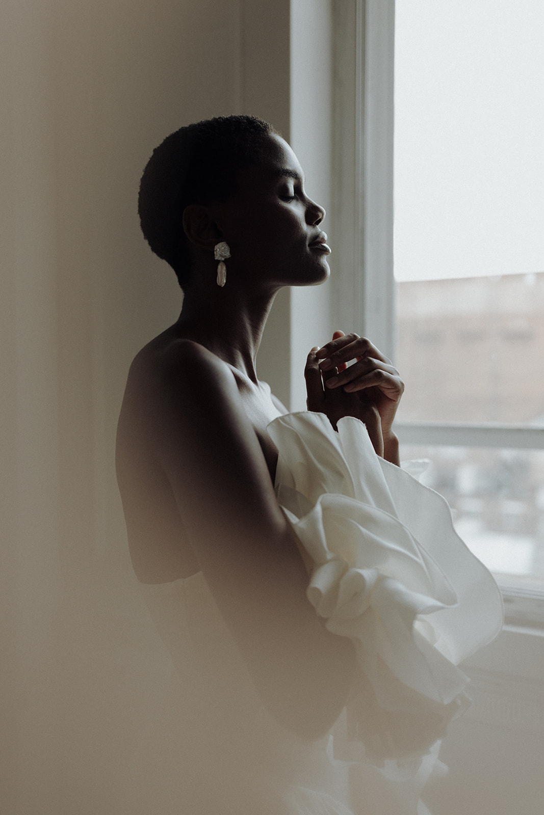 Bride in couture gown and luminous earrings, side profile at window, editorial bridal photograph.