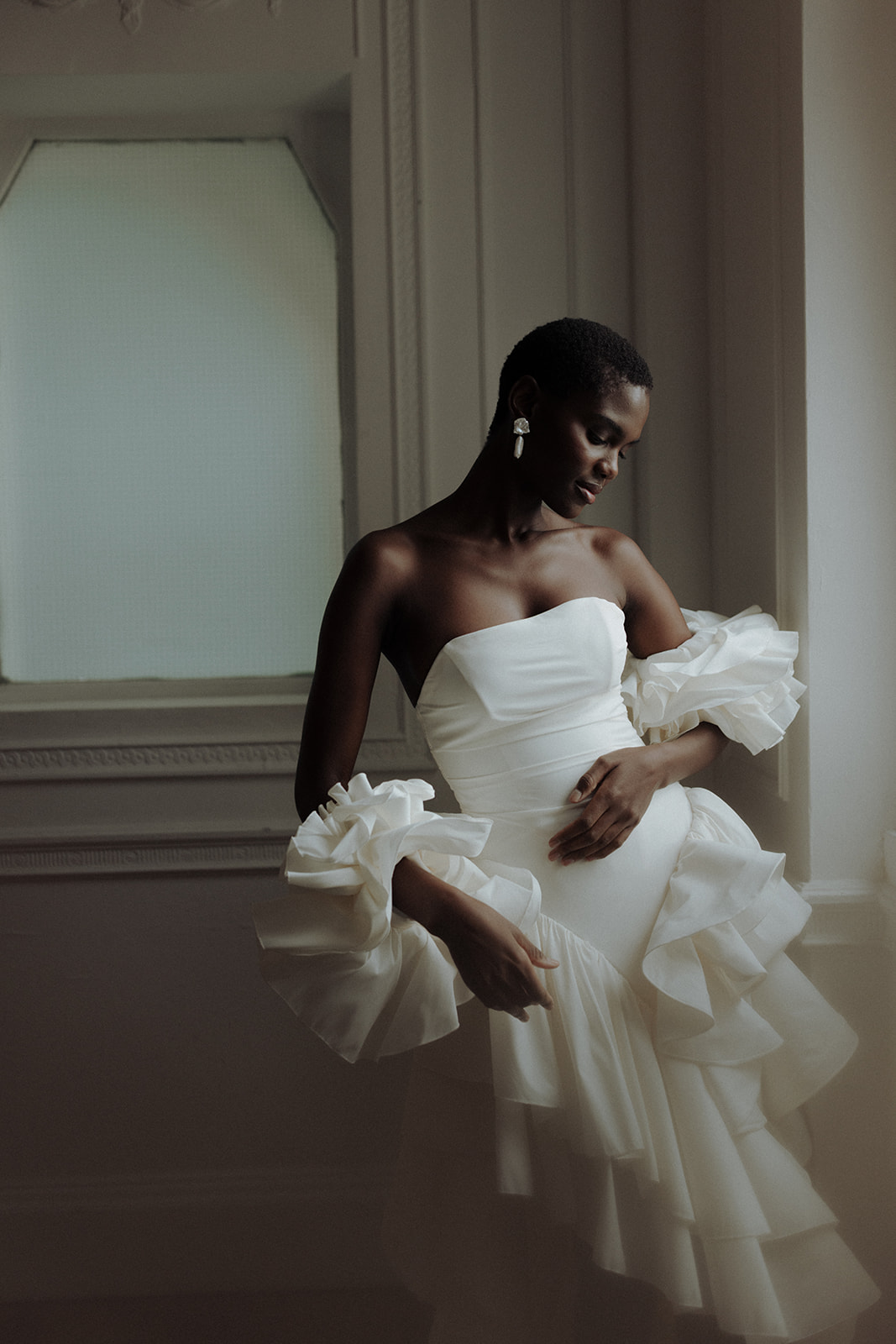 Bride in sculptural Darla Haute Couture gown standing by window, modern editorial portrait.