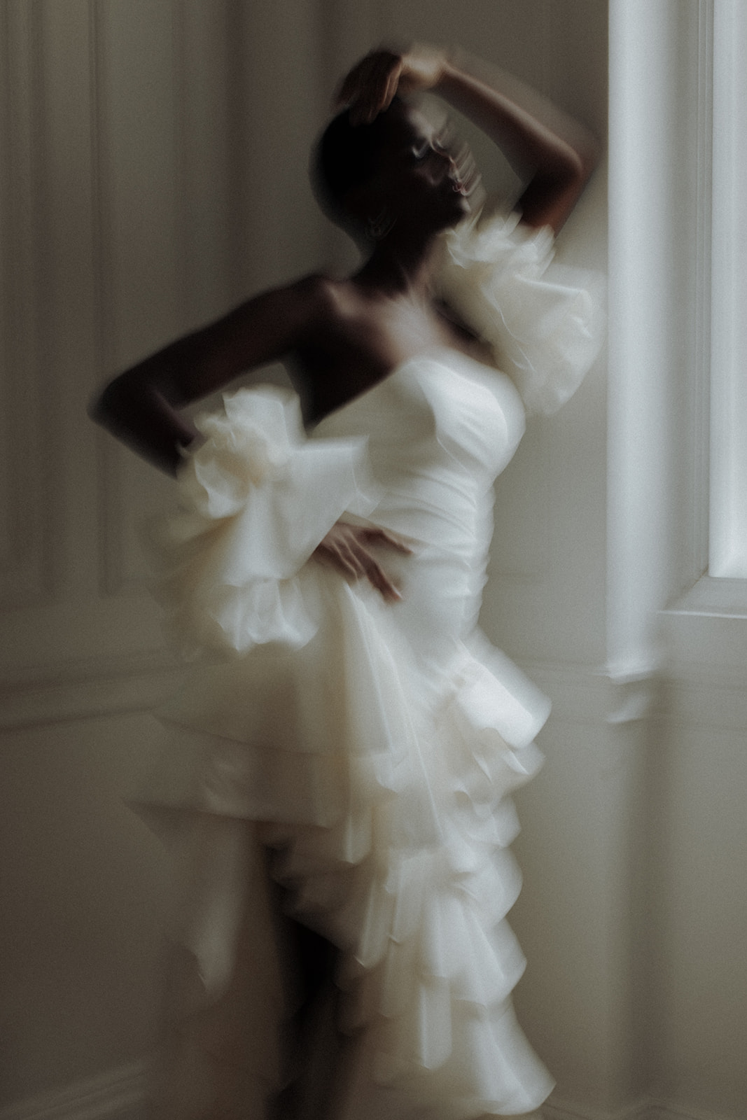 Bride in layered ruffled Darla Haute Couture gown moving by a window, blurred editorial wedding photograph.