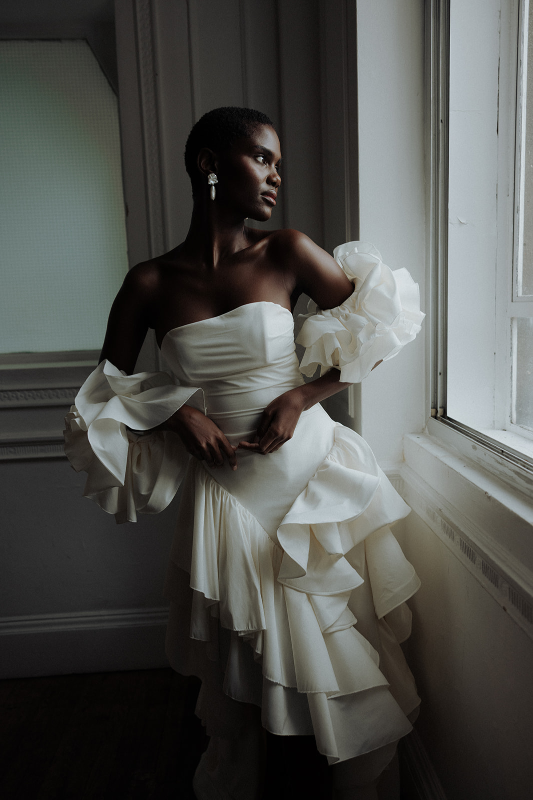 Bride in sculptural Darla Haute Couture gown standing by window, full-length editorial portrait.