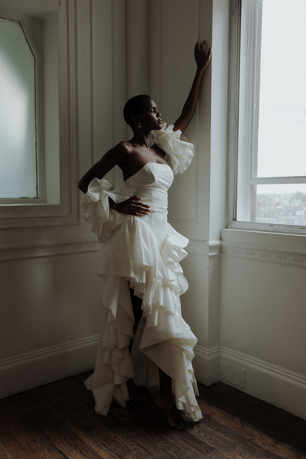 Bride in sculptural Darla Haute Couture gown standing by window, modern editorial photograph.