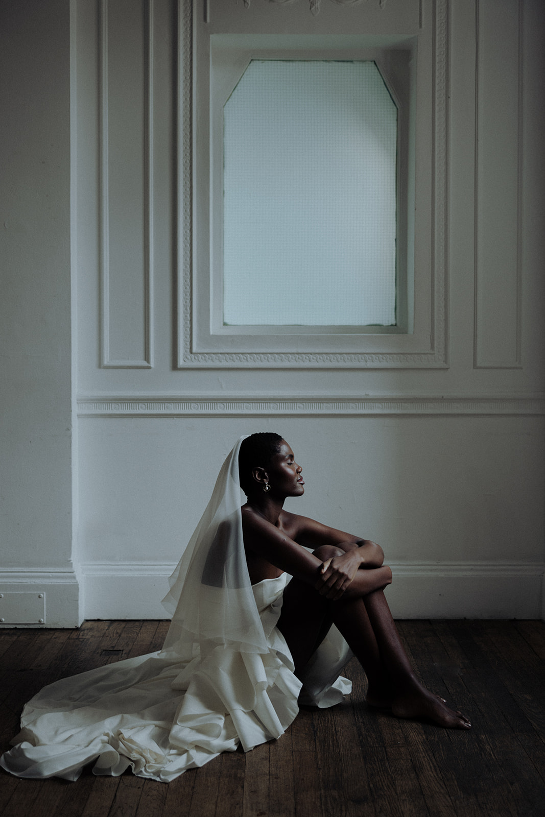 Bride in couture gown and veil seated beneath window, editorial bridal photograph.