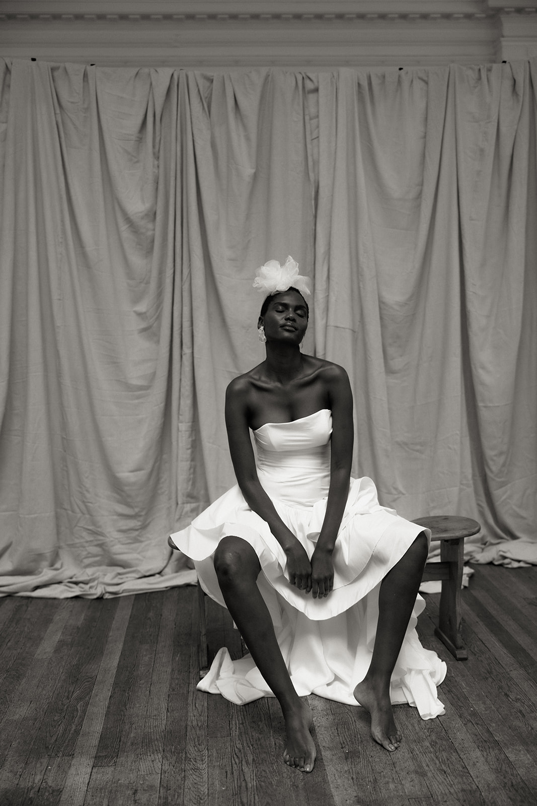 Bride wearing couture gown and headpiece, seated against draped fabric backdrop, editorial bridal portrait.