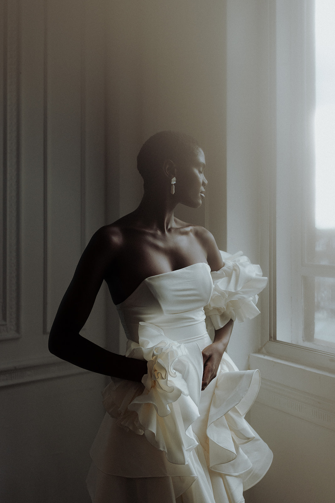 Bride in sculptural Darla Haute Couture gown gazing out window, editorial portrait.
