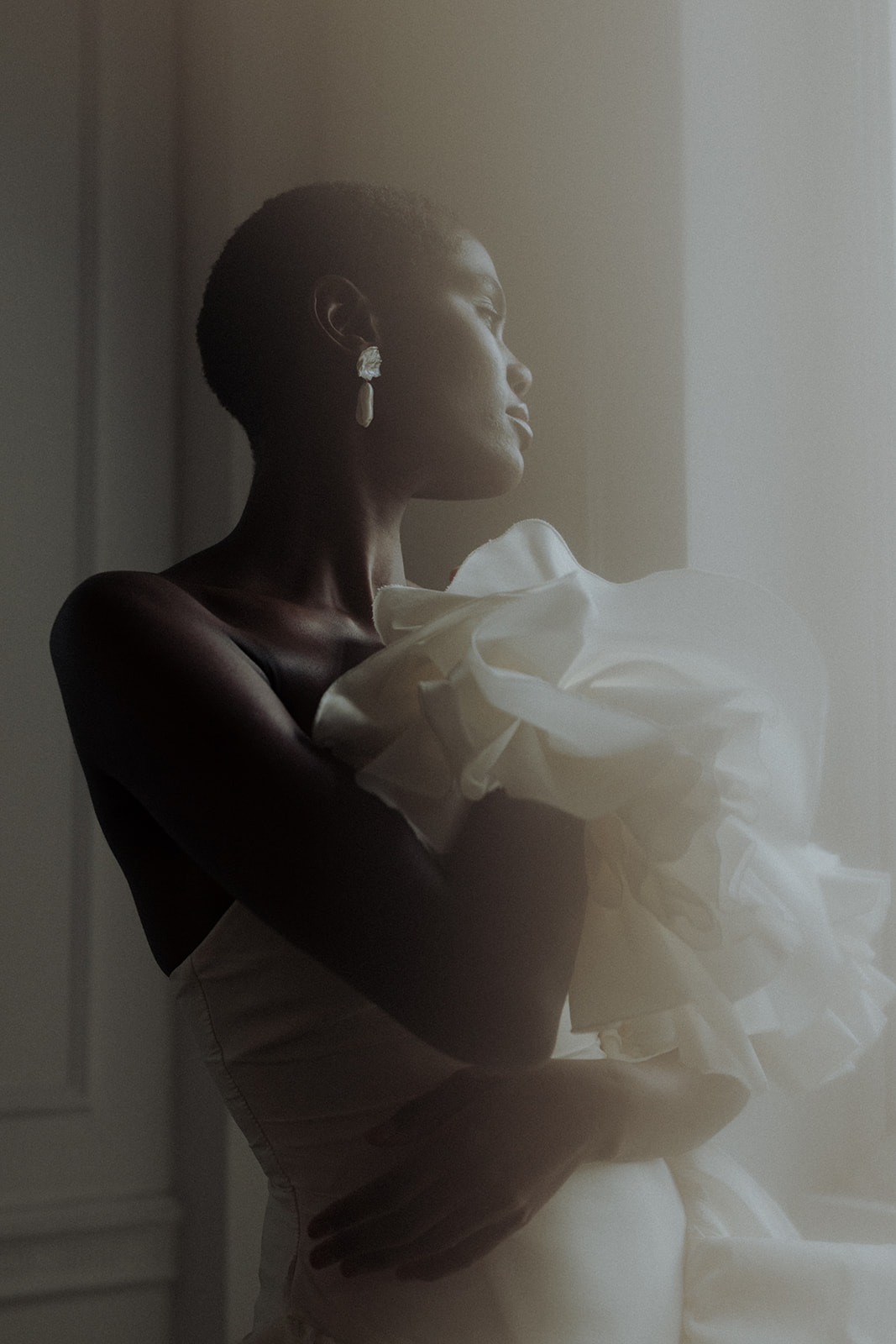 Bride in couture gown with luminous earrings, editorial portrait by window.