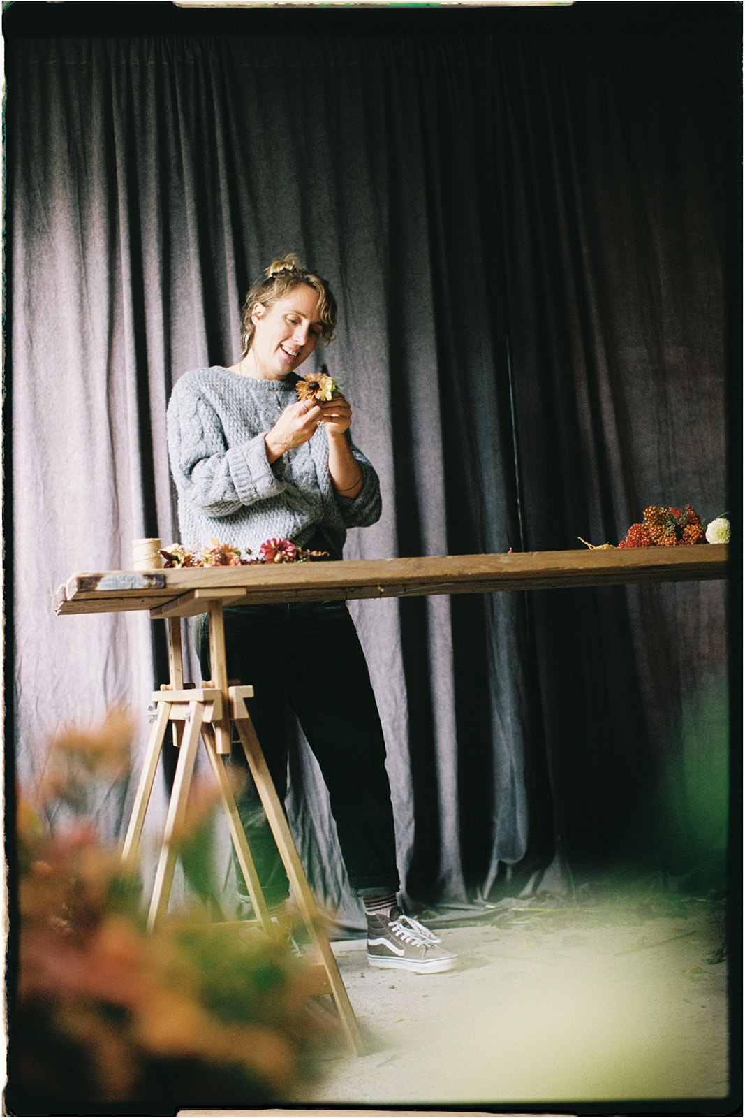 Tupelo Tree florist creating floral arrangements at a tall wooden table in soft window light, photographed on 35mm film in Scotland.